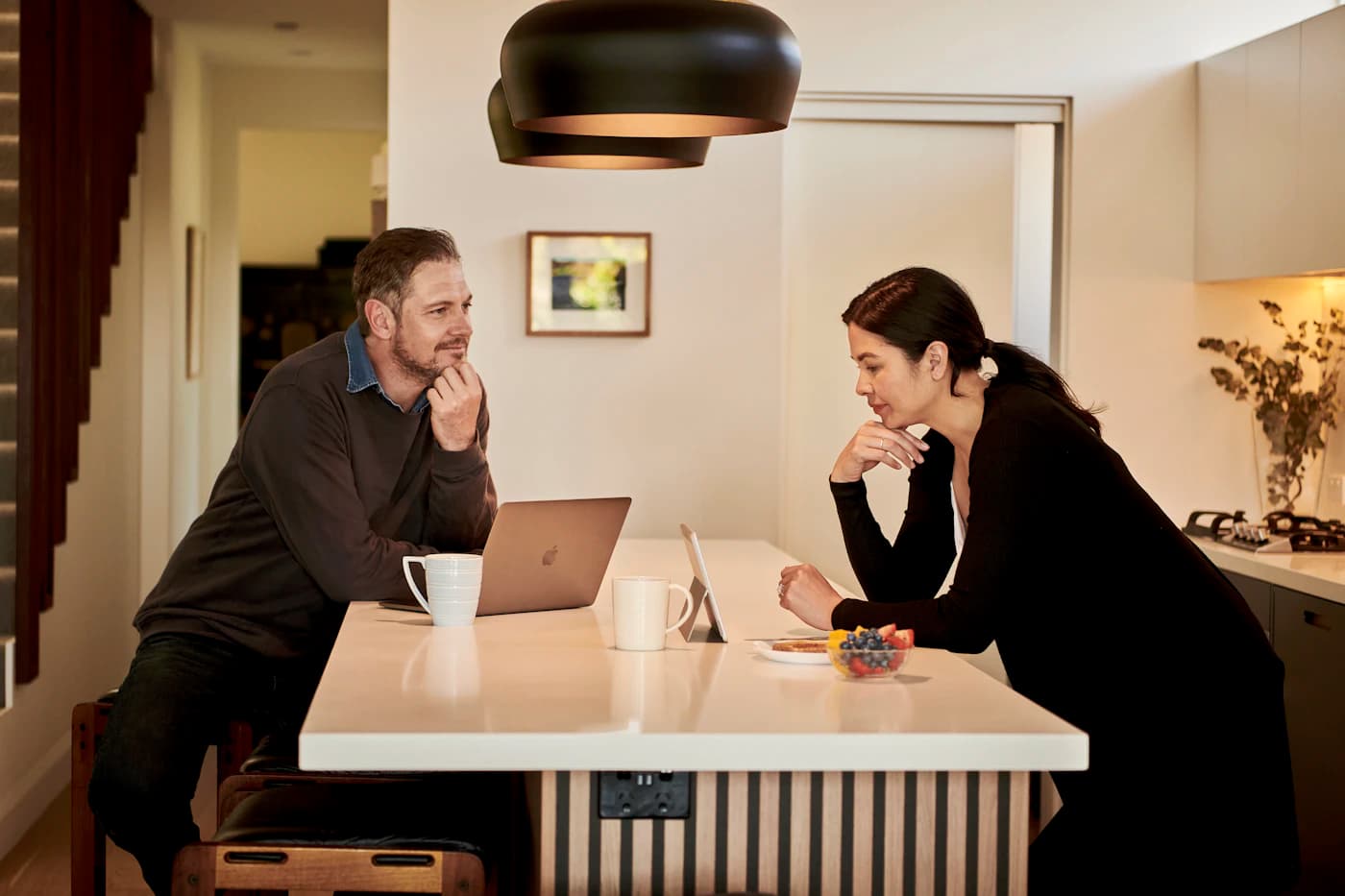 Casal conversando na bancada da cozinha, com notebook e tablet