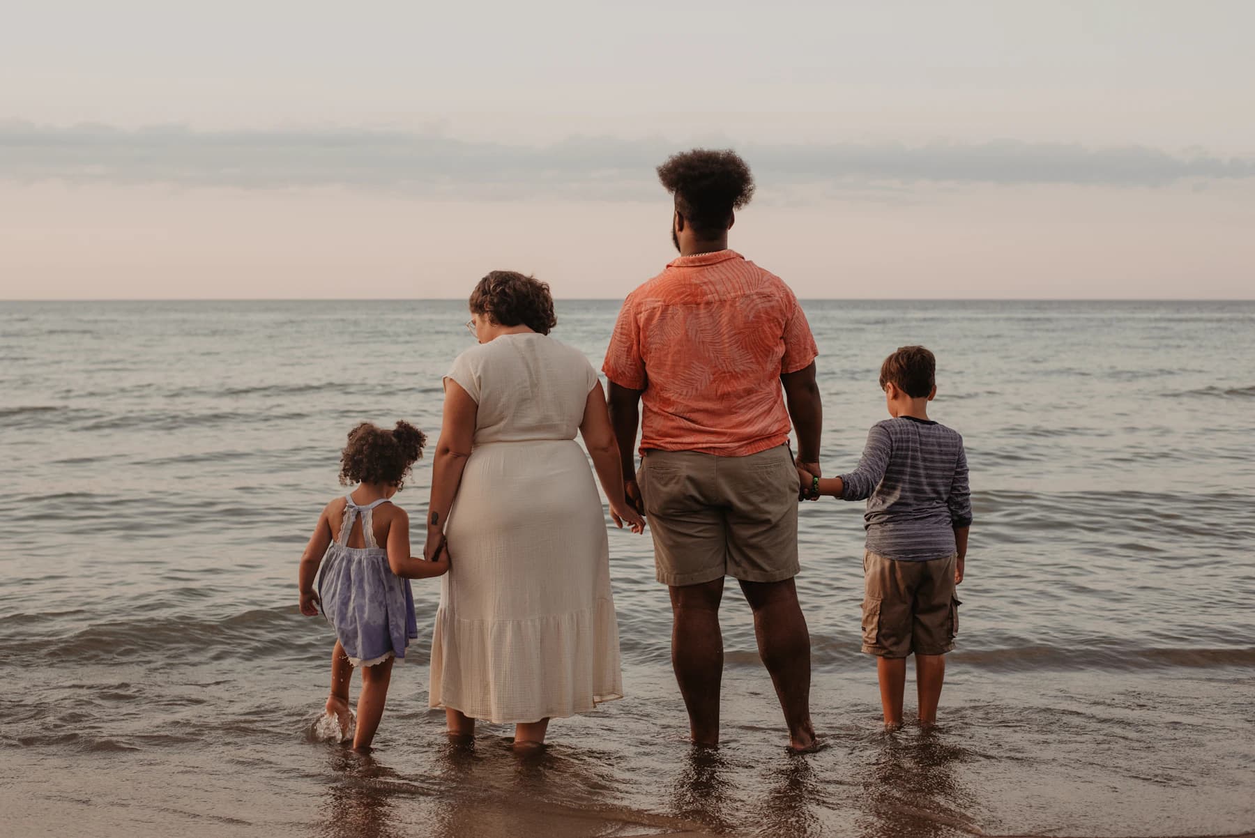 Família de mãos dadas, de costas, olhando o mar ao entardecer